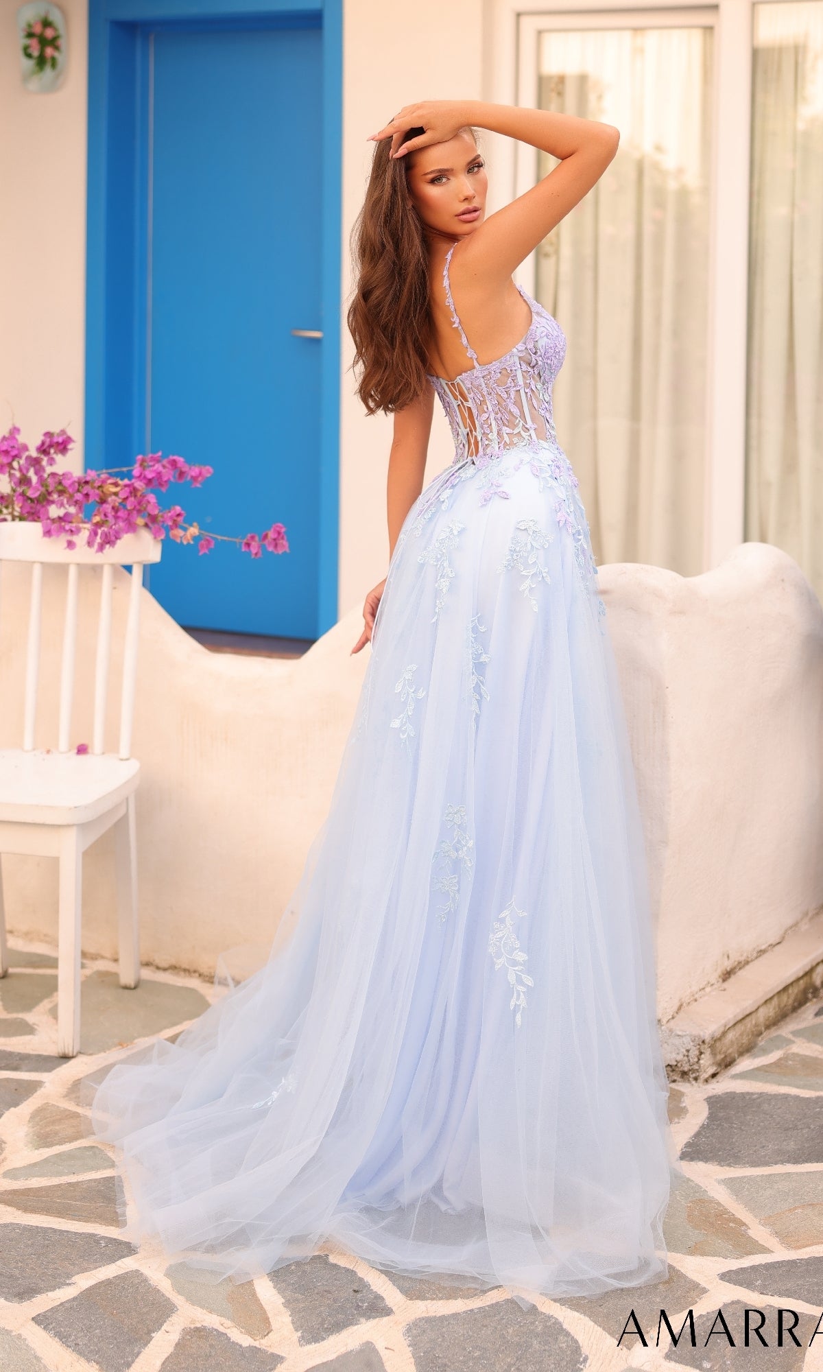 A woman in the Long Formal Dress 88115 by Amarra, a light blue floor-length A-line gown with lace embroidery, stands outdoors near a white wall and blue door, looking over her shoulder.